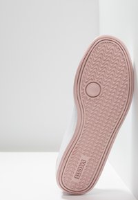 Baskets blanches avec une semelle en caoutchouc rose texturée, dotées d'une tige lisse et d'un détail de logo discret embossé sur le côté. Design plat et arrondi.