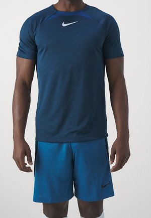Atletisk person iført en mørkeblå Nike T-shirt med korte ærmer og lysere blå Nike-shorts, stående mod en ensfarvet baggrund.