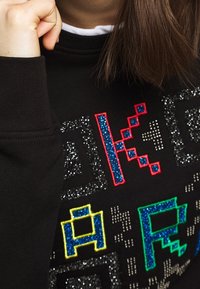 Černá mikina s texturovaným, barevným grafickým designem a třpytivými detaily. Kolo výstřih a žebrované manžety jsou viditelné.