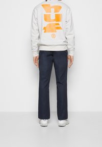 Person som bär en grå sweatshirt med en stor orange grafisk design på ryggen, marinblå byxor och vita sneakers, som står framför en enfärgad vit bakgrund.