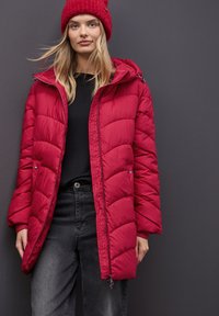 Giacca puffa rossa con cappuccio, caratterizzata da un design trapuntato, chiusura con zip e tasche laterali. Top nero e jeans completano l'outfit.
