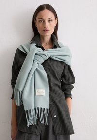 Vrouw die een donkerblauw gestreept overhemd draagt met opgerolde mouwen en een lichtblauwe sjaal om haar schouders geknoopt, staand tegen een effen witte muur.