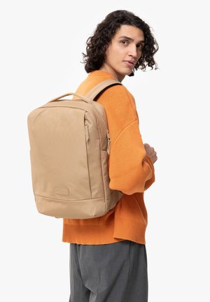 Mochila beige con un diseño rectangular, tela texturizada, bolsillo frontal y correas ajustables, llevada por una persona con un suéter naranja.