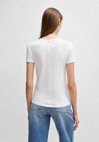 Balta trumpa rankovė t-shirt su apvalia iškirpte, pagaminta iš minkštos medžiagos, su subtiliu užpakaliniu siūlu ir mažu oranžiniu etiketu prie kaklo.