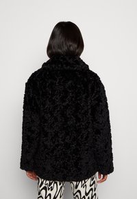 Svart faux fur-kappa med en mjuk, strukturerad yta och en bred krage. Har långa ärmar och en lös passform. Bärs över mönstrade byxor.