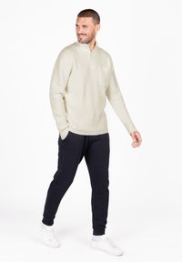 Lichtbeige wafelbrei pullover met een halve rits, voorzien van een borstzak, gecombineerd met donkerblauwe joggingsbroek en witte sneakers.
