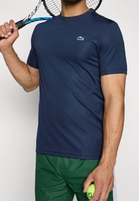 Man med marinblå skjorta och gröna shorts håller en tennisracket över axeln och en tennisboll i handen.