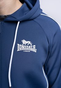 Marineblå zip-up hoodie lavet af glat stof, med et hvidt logo på brystet og kontrasterende hvide sidestriber langs ærmerne.