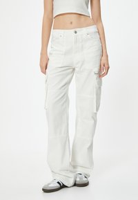 Pantaloni cargo bianchi presentano una vestibilità rilassata, numerose tasche laterali e un tessuto di cotone resistente. Il design include cuciture a vista e un piccolo dettaglio in metallo.