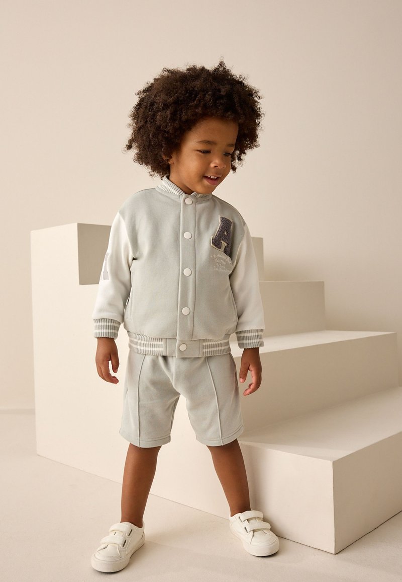 Completo grigio chiaro in due pezzi con giacca con bottoni, polsini a coste e lettera "A", abbinato a pantaloncini coordinati e scarpe slip-on bianche.