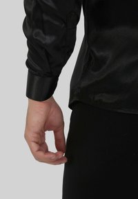 Chemise longue à manches longues en satin noir avec une texture lisse, dotée de poignets boutonnés et d'un ourlet légèrement courbé, vue de côté avec une main visible.