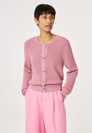 Woman with short dark hair wearing a pink knit buttoned cardigan and loose pink pants, hands in pockets, looking right.