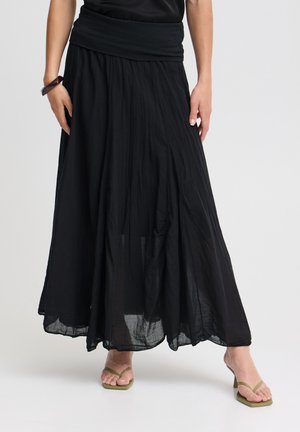 Personne portant une longue jupe noire fluide à large ceinture, des sandales à talons couleur beige et un bracelet foncé, debout sur un fond uni.