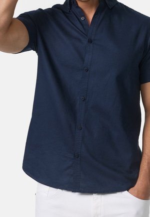 Homme portant une chemise à manches courtes bleu marine boutonnée, une main dans la poche et l'autre levée, sur un fond uni.