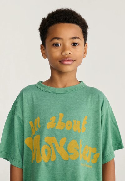 Jeune garçon aux cheveux courts et bouclés portant un t-shirt vert avec un texte jaune indiquant « "All about Monsters" » sur un fond clair uni.
