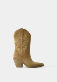 Beżowy zamszowy kowbojski but z zaostrzonym noskiem i profilowaną cholewką. Ozdobiony białymi haftowanymi wzorami w kształcie wirów oraz posiadający piętę typu stack.