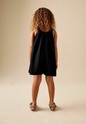 Kind mit lockigem Haar, das ein ärmelloses schwarzes Kleid und Sandalen mit Leopardenmuster trägt, steht vor einem beigen Hintergrund.