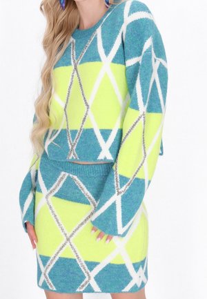 Femme portant un ensemble pull et jupe à motif losanges turquoise, jaune et blanc, avec de longs cheveux blonds ondulés et des boucles d’oreilles violettes.