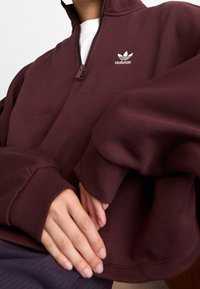 Burgundyröd sweatshirt med halv dragkedja, tillverkad av mjukt material med hög krage, med en vit broderad Adidas-logotyp på bröstet och avslappnade ärmar.