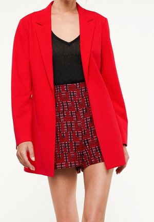 Mujer con una chaqueta roja brillante sobre una camiseta negra y pantalones cortos rojos con textura y estampado, sobre un fondo blanco.
