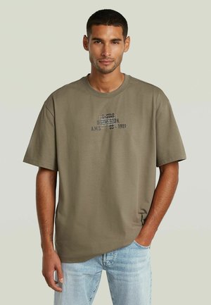 Junger Mann mit kurzen dunklen Haaren, trägt ein olivgrünes übergroßes T-Shirt mit Text und hellblaue Jeans, steht mit einer Hand in der Tasche.