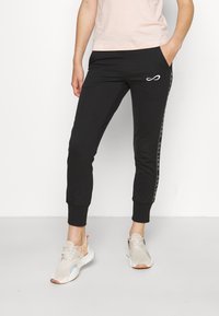 Svarta joggerbyxor med tapered passform, vit logodetalj, sidostråkar med text och elastiska muddar. Burit med ljusa sneakers.