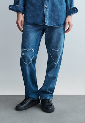 Personne portant un jean en denim bleu avec des patchs en forme de cœur usés aux deux genoux, associée à des baskets noires et une chemise en denim assortie.
