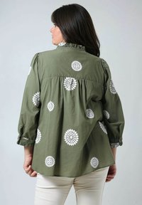 Olijfgroene blouse met witte bloemenborduursels. Kenmerkt zich door een gerimpelde kraag en driekwartmouwen, gemaakt van lichte stof.