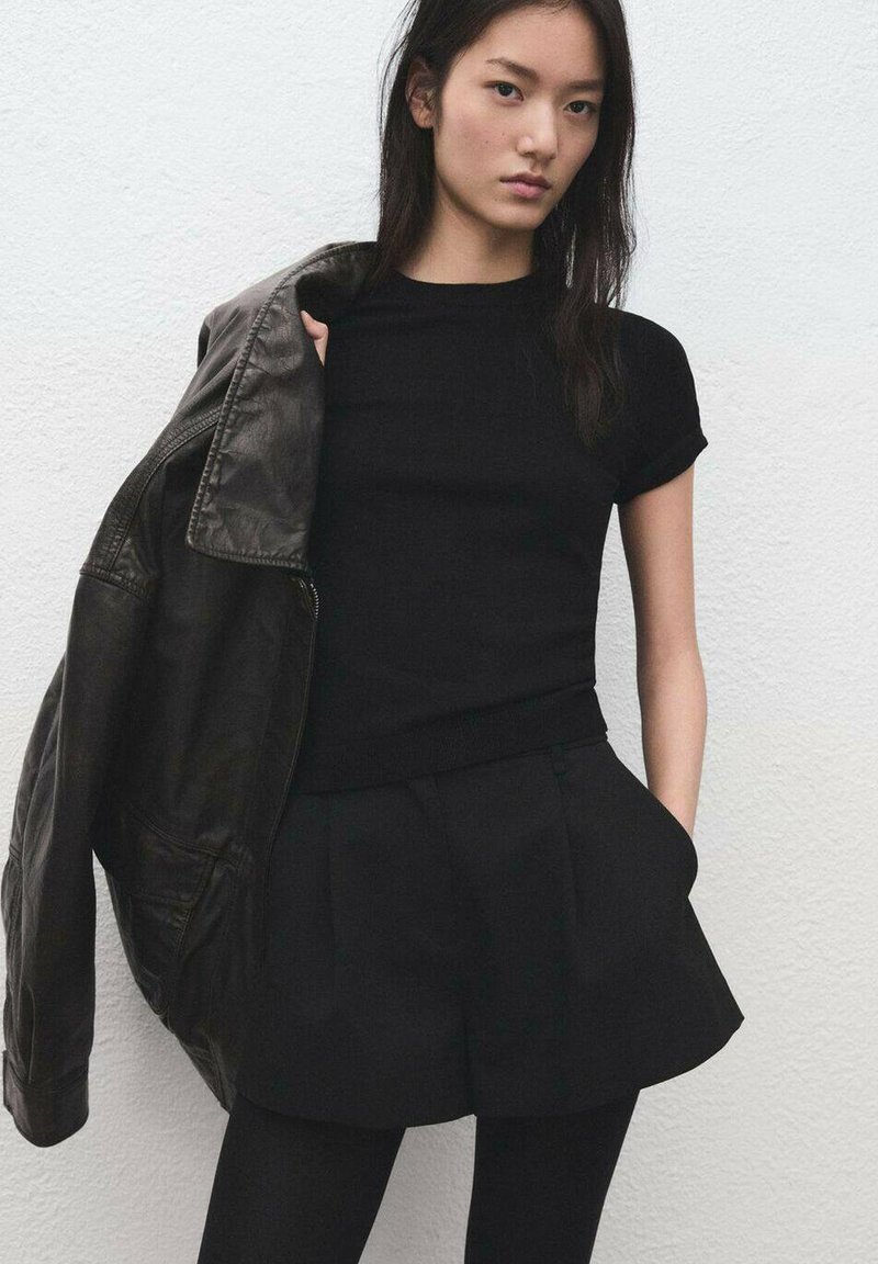 T-shirt noir ajusté associé à une veste en cuir noire. Le modèle porte un short plissé noir et des collants opaques noirs sur un fond blanc.