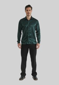 Chemise en satin vert foncé à manches longues avec un devant boutonné, un col pointu et une coupe ajustée, associée à un pantalon et des chaussures noirs.