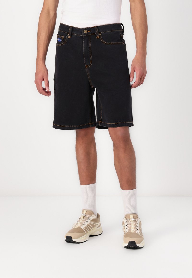 Sorte denim shorts med orange syninger, forlommer og en sidelomme, båret med beige sneakers og hvide ankelsokker.