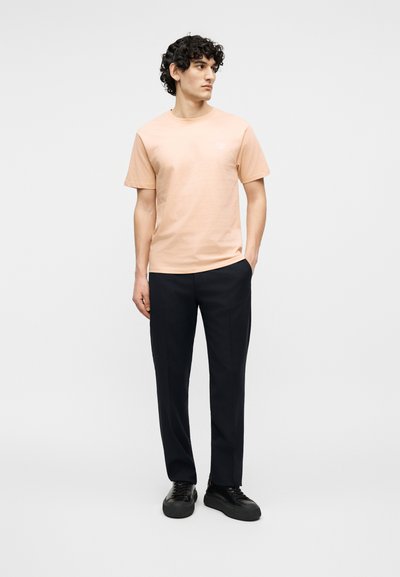 Νεαρός άνδρας με σγουρά μαλλιά, που φοράει ροδακινί κοντομάνικη μπλούζα, μαύρο παντελόνι και μαύρα αθλητικά παπούτσια, στέκεται μπροστά σε απλό ανοιχτό φόντο.