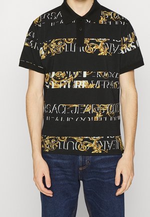 Man draagt een zwart poloshirt met korte mouwen met gouden barokprint en witte gefragmenteerde tekst, gecombineerd met donkerblauwe spijkerbroek, staand tegen een witte achtergrond.