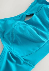 Sutien sport de culoare turquoise, realizat dintr-un material neted și elastic. Prezintă un decolteu rotund, cusături și o detaliu tăiat la spate.