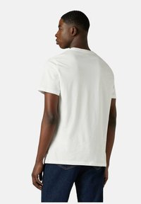 Mand iført en enkel hvid kortærmet t-shirt og mørkeblå jeans, vendt væk mod en ensfarvet lys grå baggrund.