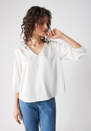 White, v-neck blouse with three-quarter sleeves, crafted from a smooth fabric. Paired with blue denim jeans. Simple design, no notable patterns.