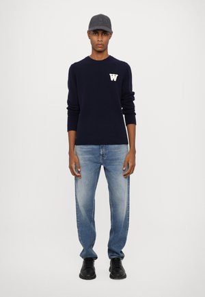 Jersey azul marino con una letra "W" blanca en el pecho, vaqueros azul claro con un aspecto desgastado y zapatos negros. Gorro simple con un acento de logo.