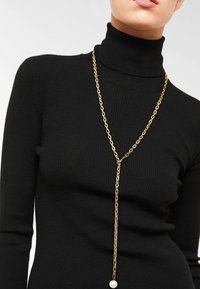 Guldkæde med en lang dråbe og en perleaccent, båret over en sort ribbet turtleneck. En enkel og elegant design.
