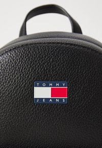 Schwarzer, strukturierten Leder-Rucksack mit einem markanten rechteckigen Logo, auf dem "TOMMY JEANS" in Blau, Rot und Weiß steht. Runde Form mit einem oberen Griff.