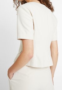 Femme portant un blazer crème ajusté à manches courtes et une jupe assortie, une main dans la poche, vue de dos et de côté.