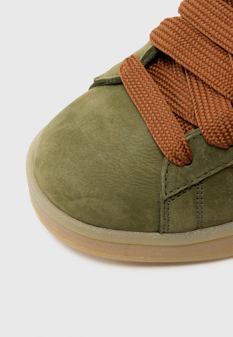 Olivgrüner Wildleder-Sneaker mit rund zulaufender Spitze, strukturierten braunen Schnürsenkeln und hellbrauner Gummisohle. Mit gesteppten Details an der Seite.