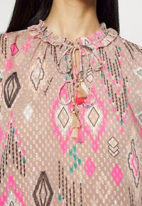 Gros plan sur un décolleté avec un chemisier rose-beige à motifs, doté d'un col volanté et de liens à franges en beige et rose vif.