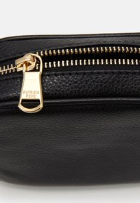 Černá texturovaná kožená taštička s zlatým zipem a gravírovaným brandingem. Detailní pohled se zaměřuje na zip a texturu materiálu.