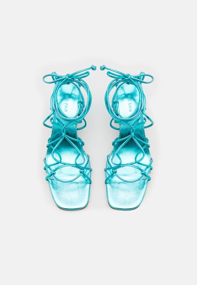 Ένα ζευγάρι τιρκουάζ σандάλια με τετράγωνη μύτη, λεπτές ρίγες και γυαλιστερή σατέν φινίρισμα με πολλές λεπτομέρειες δέσμευσης.