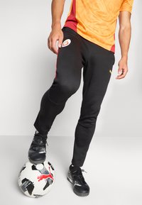 Sorte træningsbukser med røde side striber, logo på låret, parret med en orange og rød struktureret skjorte, stående på en mønstret fodbold.