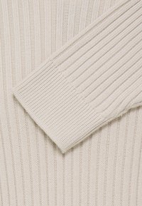 Gros plan d'un tissu de pull tricoté crème montrant une texture côtelée et un revers de manche plié.