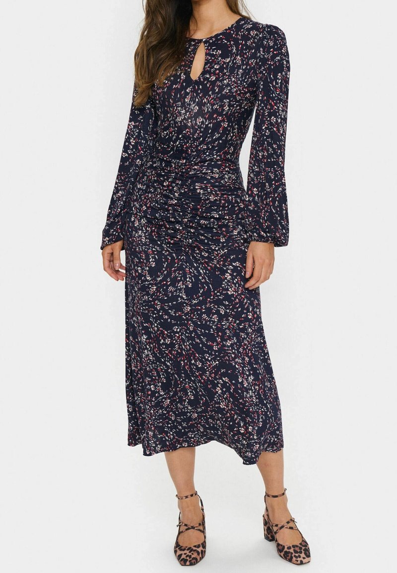 Femme portant une robe midi bleu marine à manches longues, taille froncée, motif floral abstrait blanc et rouge, associée à des escarpins à bride cheville imprimé léopard.