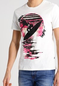 Vit bomull t-shirt med en grafisk silhuett av en surfare mot en rosa och svart cirkulär solnedgångsdesign. Kort ärm, rund hals.