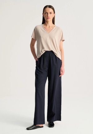 Kobieta stojąca, ubrana w beżowy top z krótkim rękawem i dekoltem w serek, czarne spodnie z szerokimi nogawkami, czarne wsuwane buty, ręce w stanie odprężenia, neutralne tło.