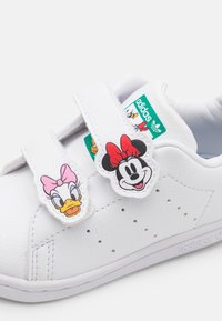 Bílá teniska s dvěma suchými zipy, s nášivkami postaviček Daisy Duck a Minnie Mouse. Vyrobená z hladkého syntetického materiálu s logem.